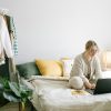 A woman working from home on a laptop in a cozy, stylish bedroom setting.